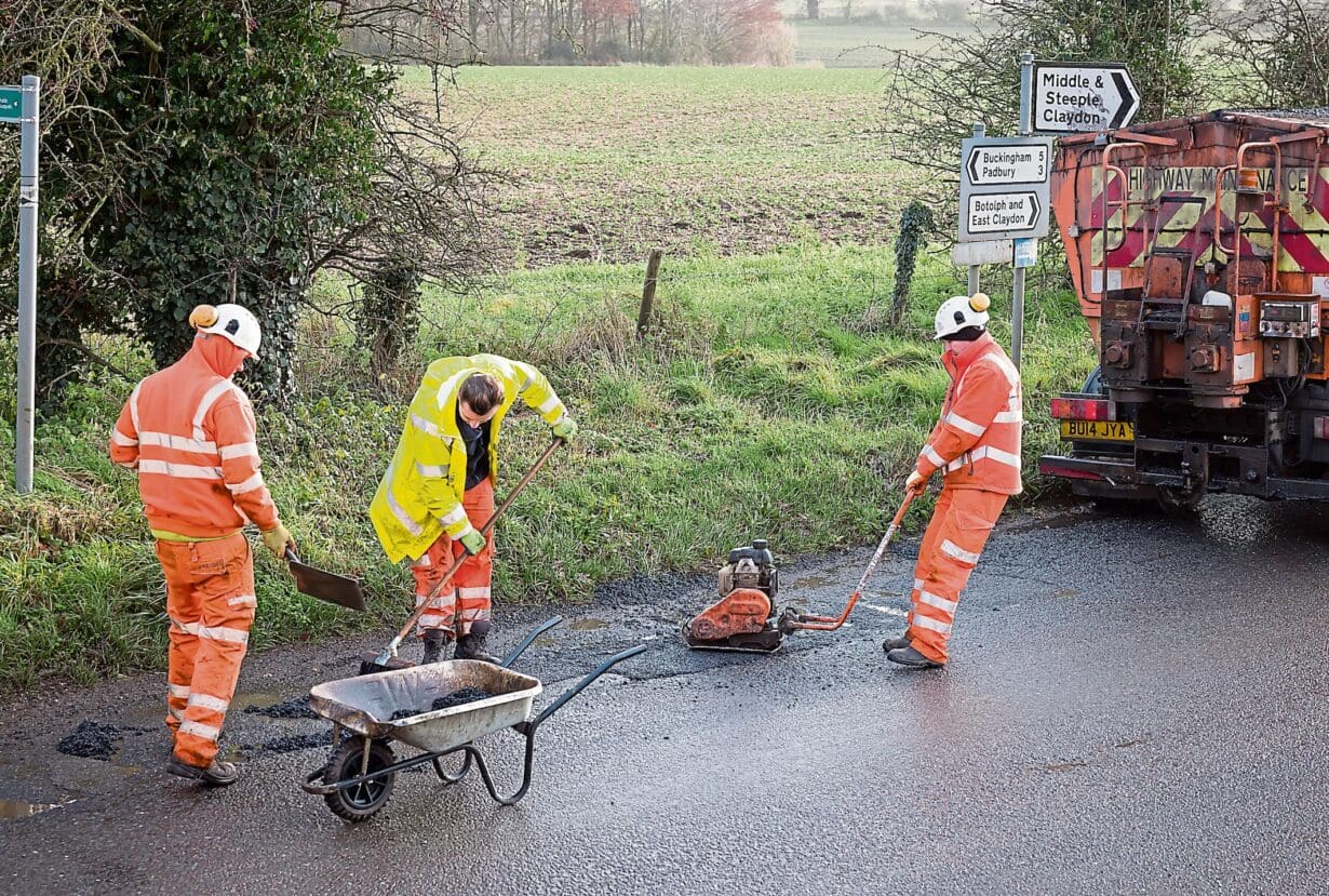 Buckingham, UK - December 14, 2020. Road works, road crew team, men wearing high visibility hi vis orange clothing. Repairing potholes in a rural country road